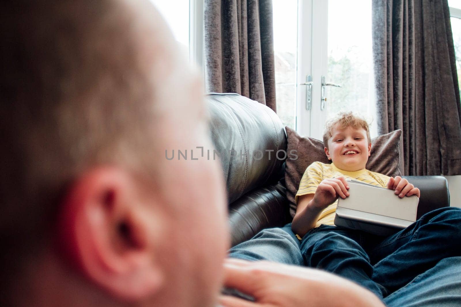 Little boy and his father are talking while lying on the sofa together. The little boy is holding a digital tablet.