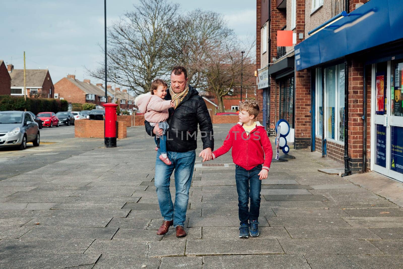 Mature man is taking his children to the local shop with him. He is holding his sons hand and carrying his baby daughter on his hip.