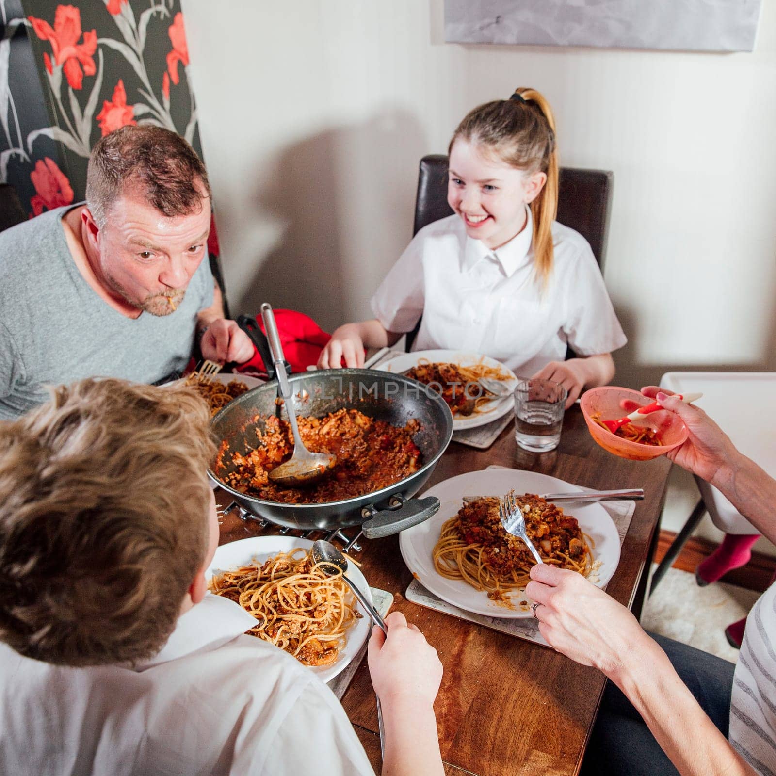 British family enjoying a spagetti bolognese together at home. The children are laughing as their father has spagetti hanging out his mouth.