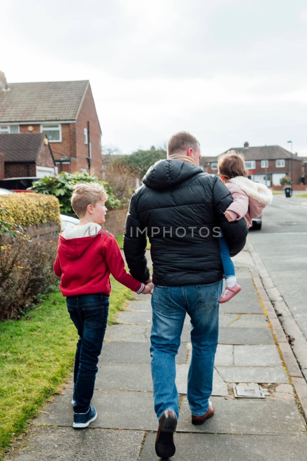 Rear view of a mature man walking down the street with his children. He is holding his sons hand and carrying his baby daughter.