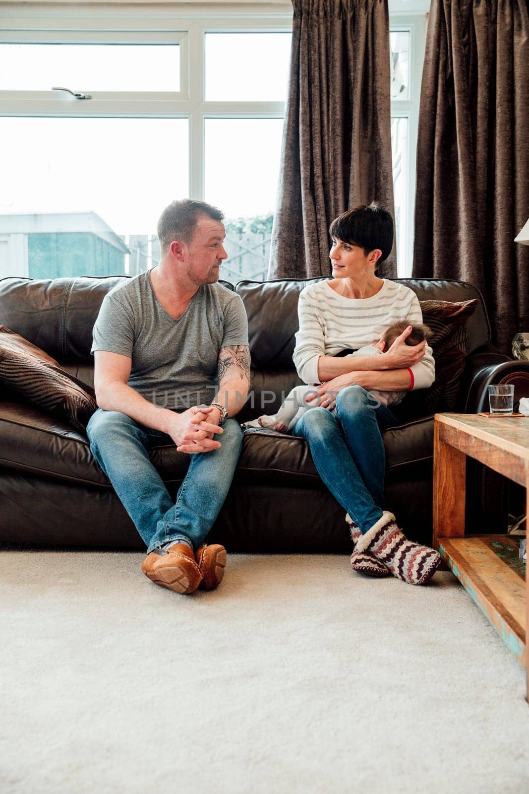Mid adult couple are chatting on the sofa in the living room of their home while the woman is breastfeeding their baby daughter.