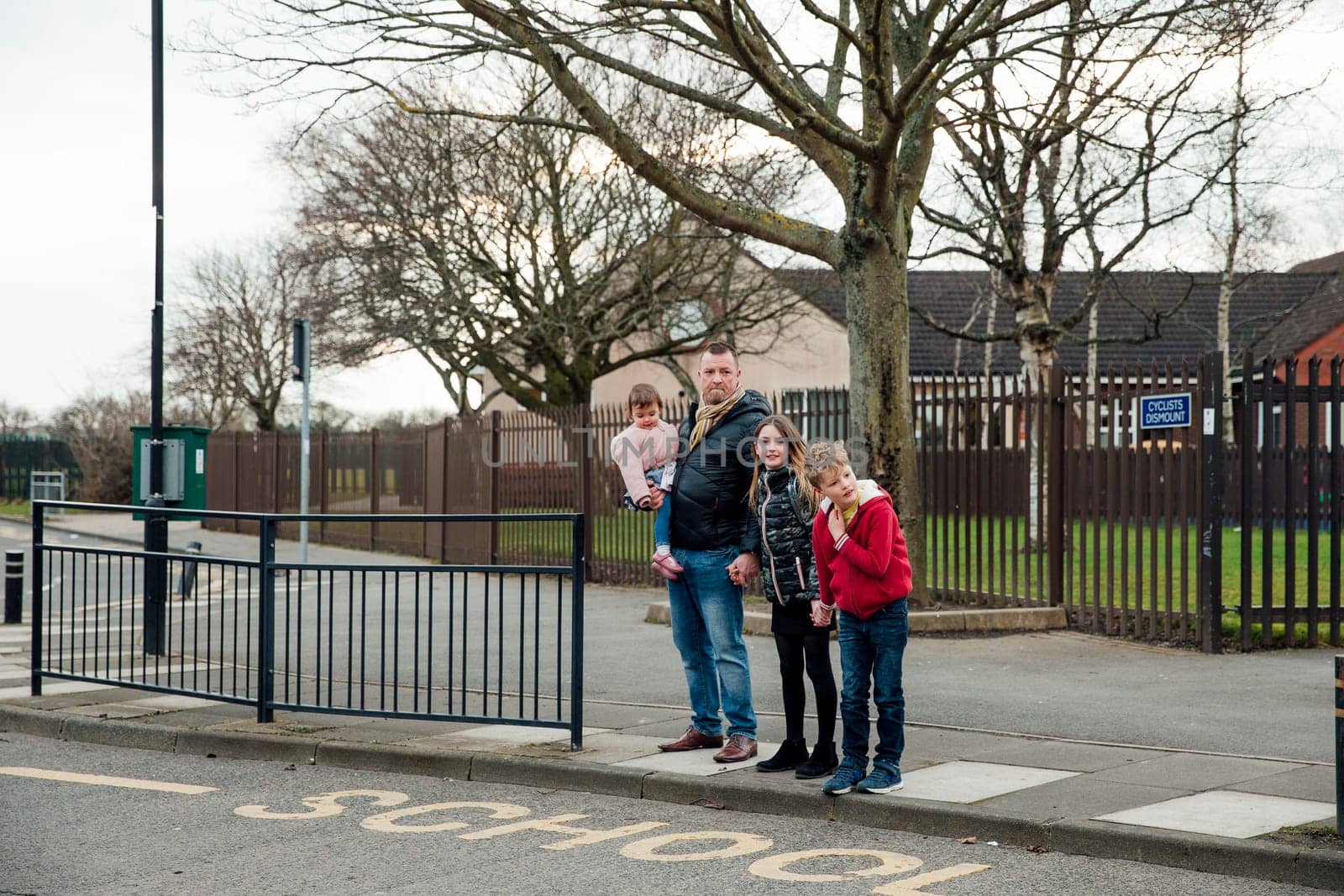 Mature man and his three children are waiting to cross the road. They have picked the eldest daughter up from school and are going home.