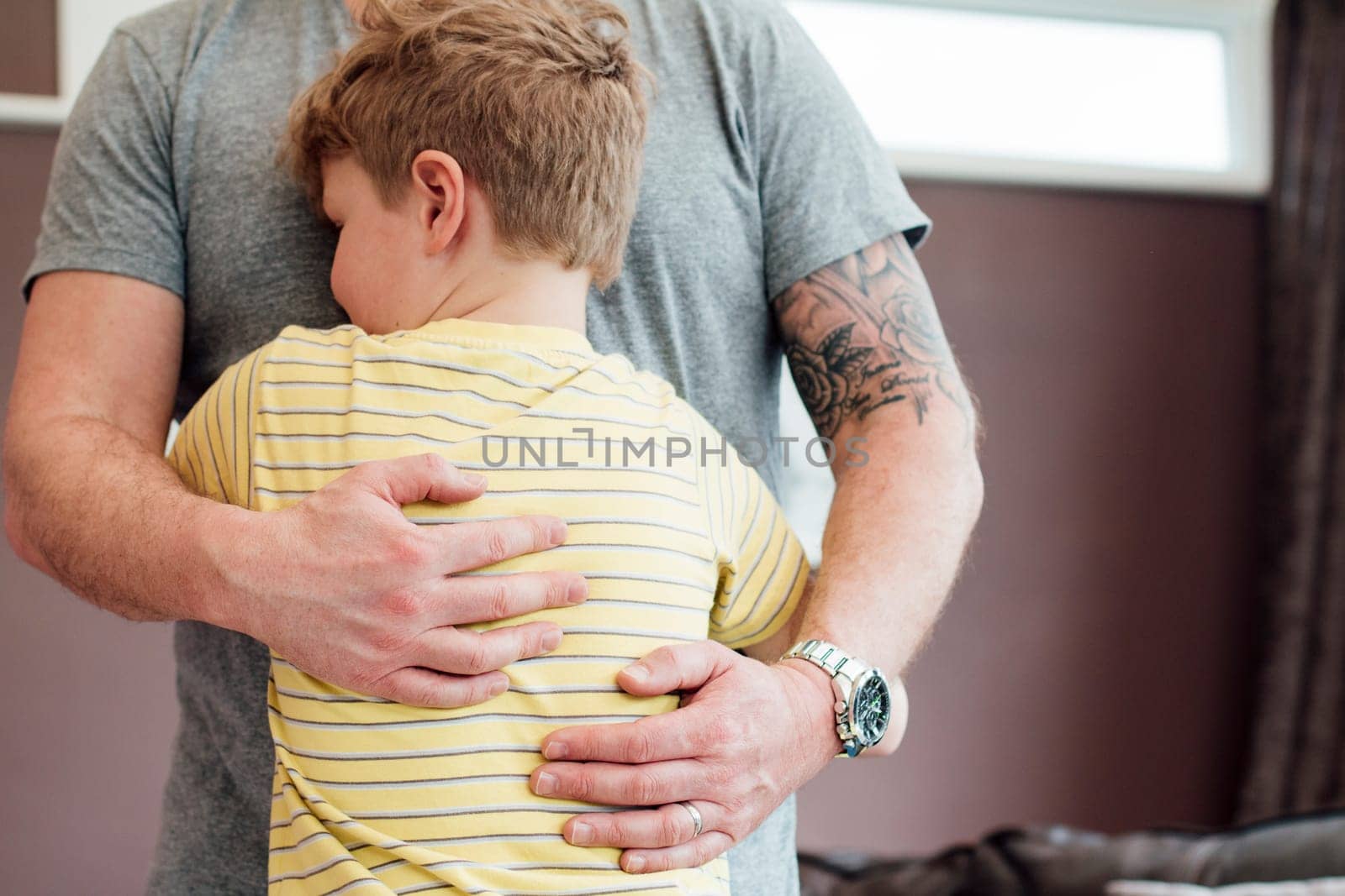 Little boy is cuddling his father at home. He has his arms round his fathers torso and the father is holding his son close to him.