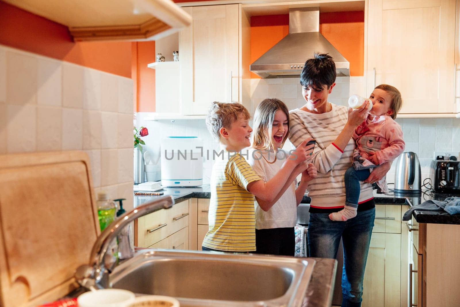Girl and her brother are showing their mum something on their smartphone. They are all laughing and the mother has the baby on her hip, feeding her at the same time.
