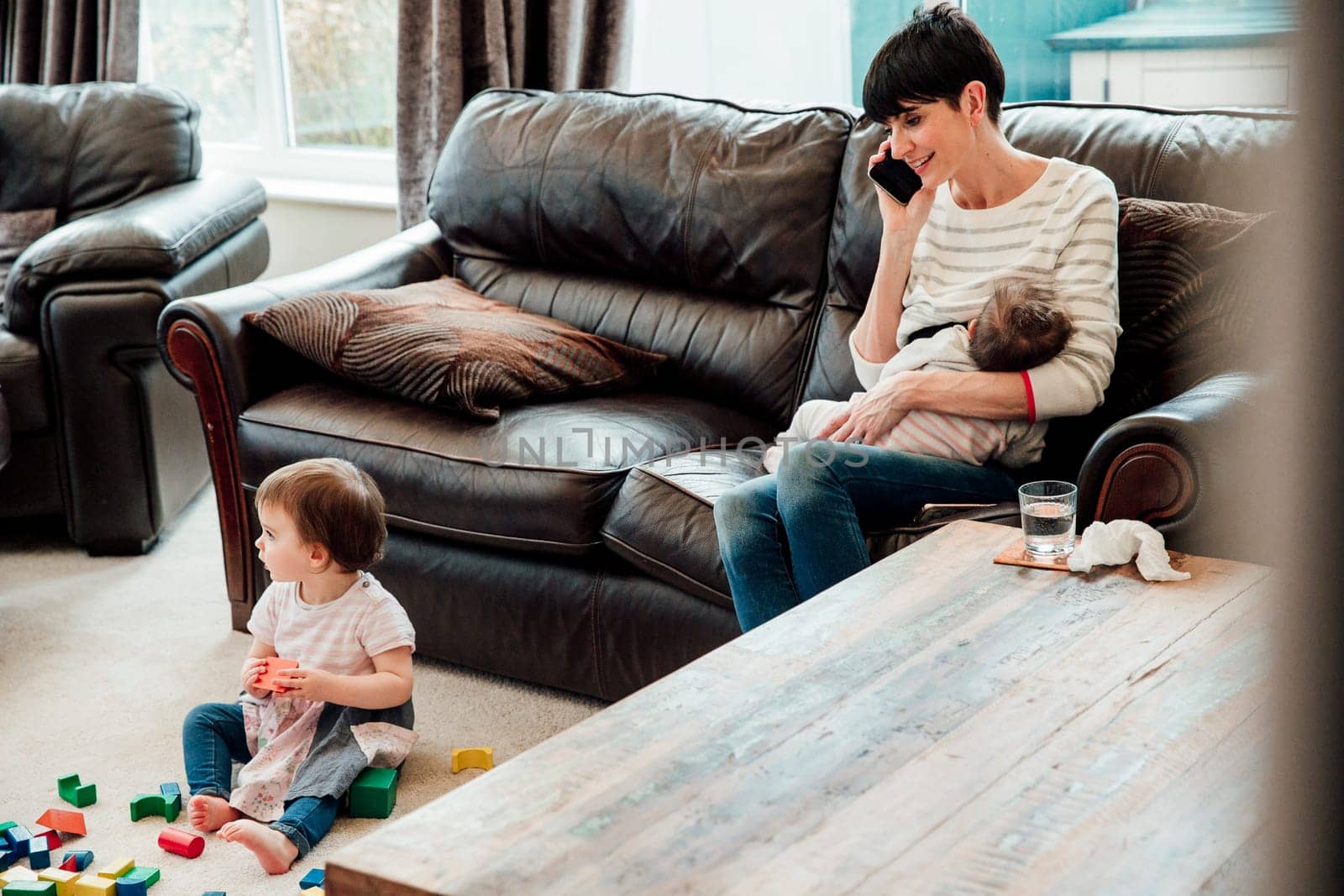Mid adult mother is multitasking at home. she is sitting on the sofa in the living room, breast feeding her youngest baby and watching her older daughter play while talking on the phone.