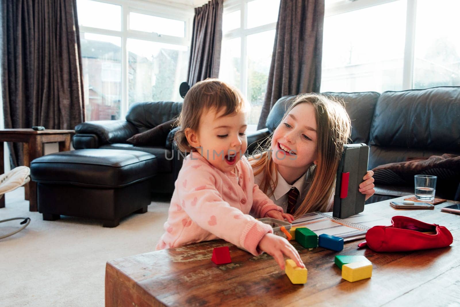 Little girl is distracting her older sister while she is trying to study at home. They are playing with toy blocks together.