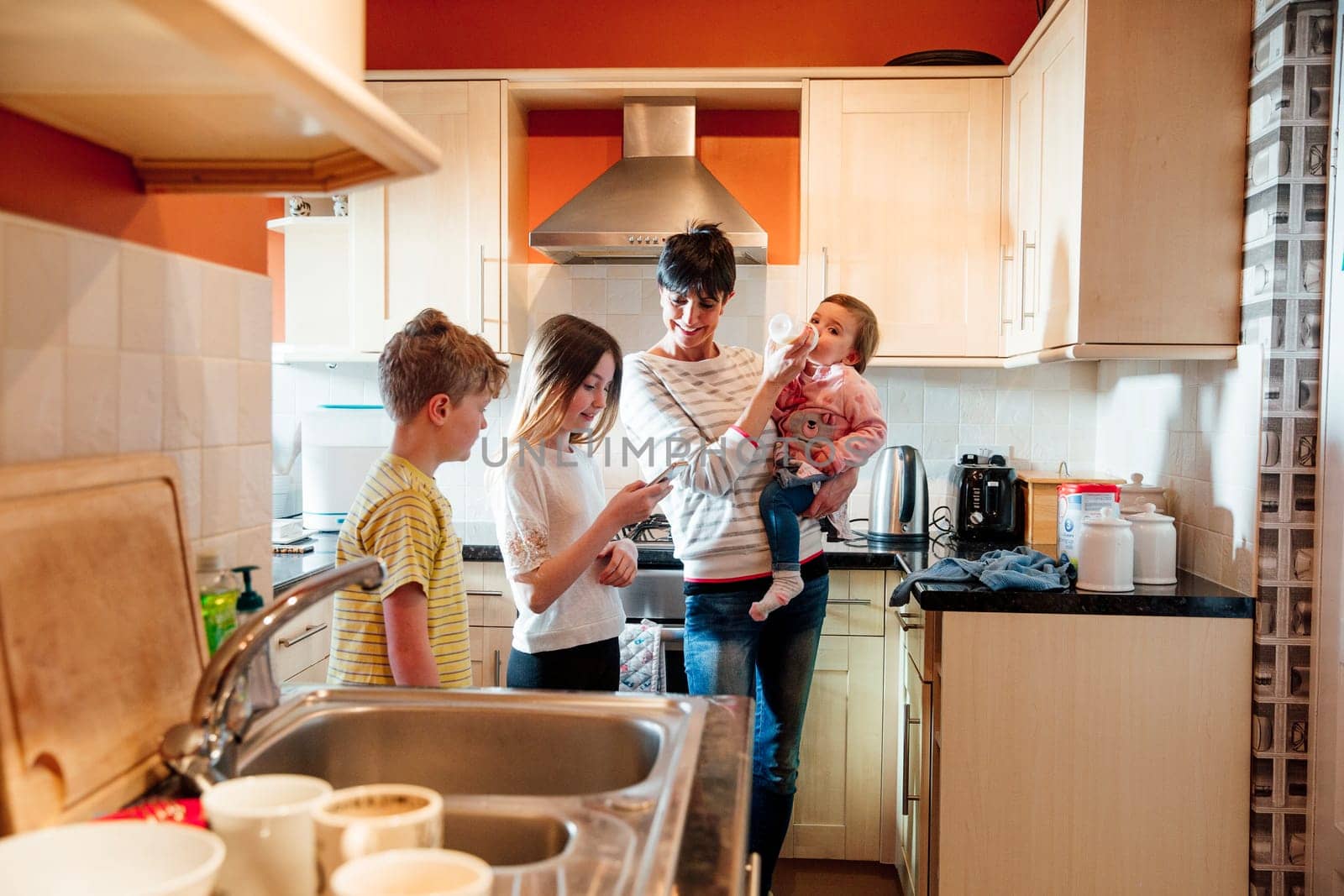 Multitasking mum is tending to all of her children at once in the kitchen of their home. She is feeding the baby a bottle while talking to her eldest children and looking at their smart phone.