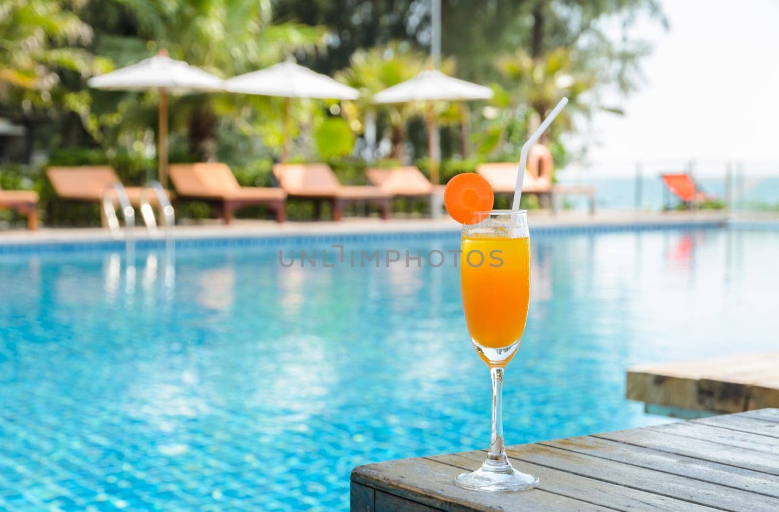 Orange juice with carrot slice in cocktail glass on wooden table at outdoor swimming pool, summer tropical holiday concept