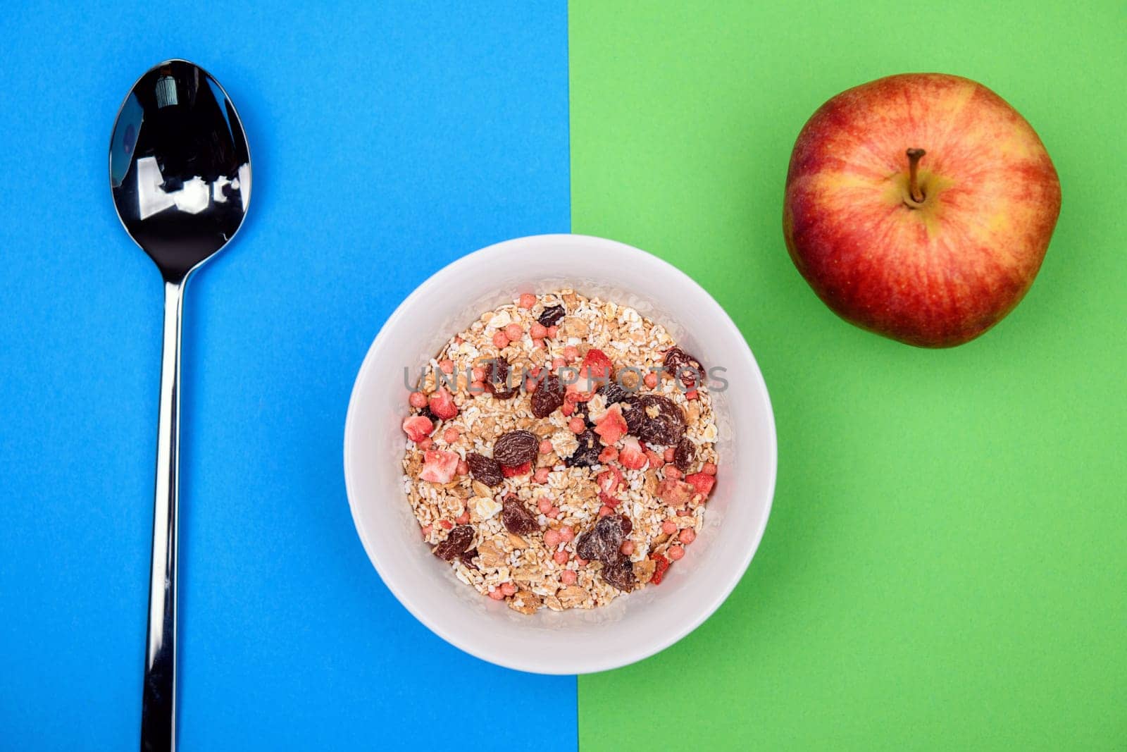 muesli in white bowl on blue and green ground with spoon and apple from above
