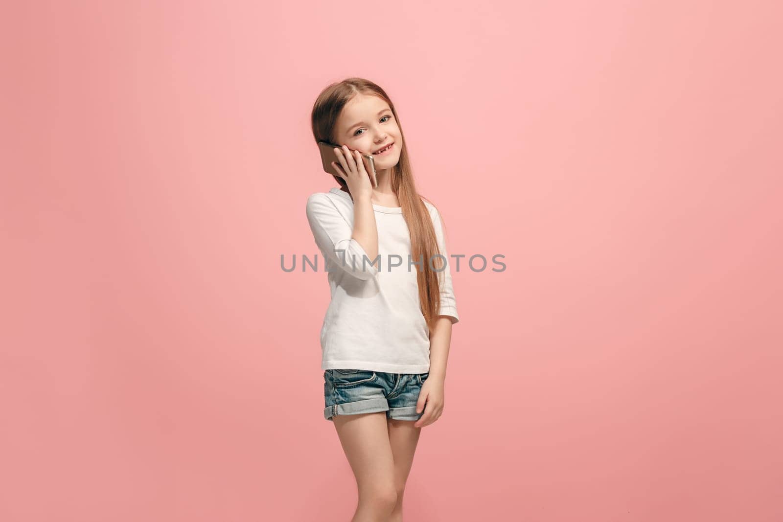 Happy teen girl standing, smiling with mobile phone over trendy pink studio background. Beautiful female half-length portrait. Human emotions, facial expression concept.