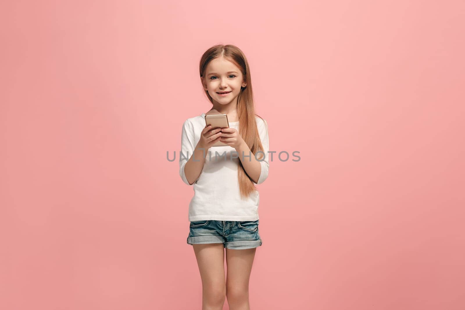 Happy teen girl standing, smiling with mobile phone over trendy pink studio background. Beautiful female half-length portrait. Human emotions, facial expression concept.