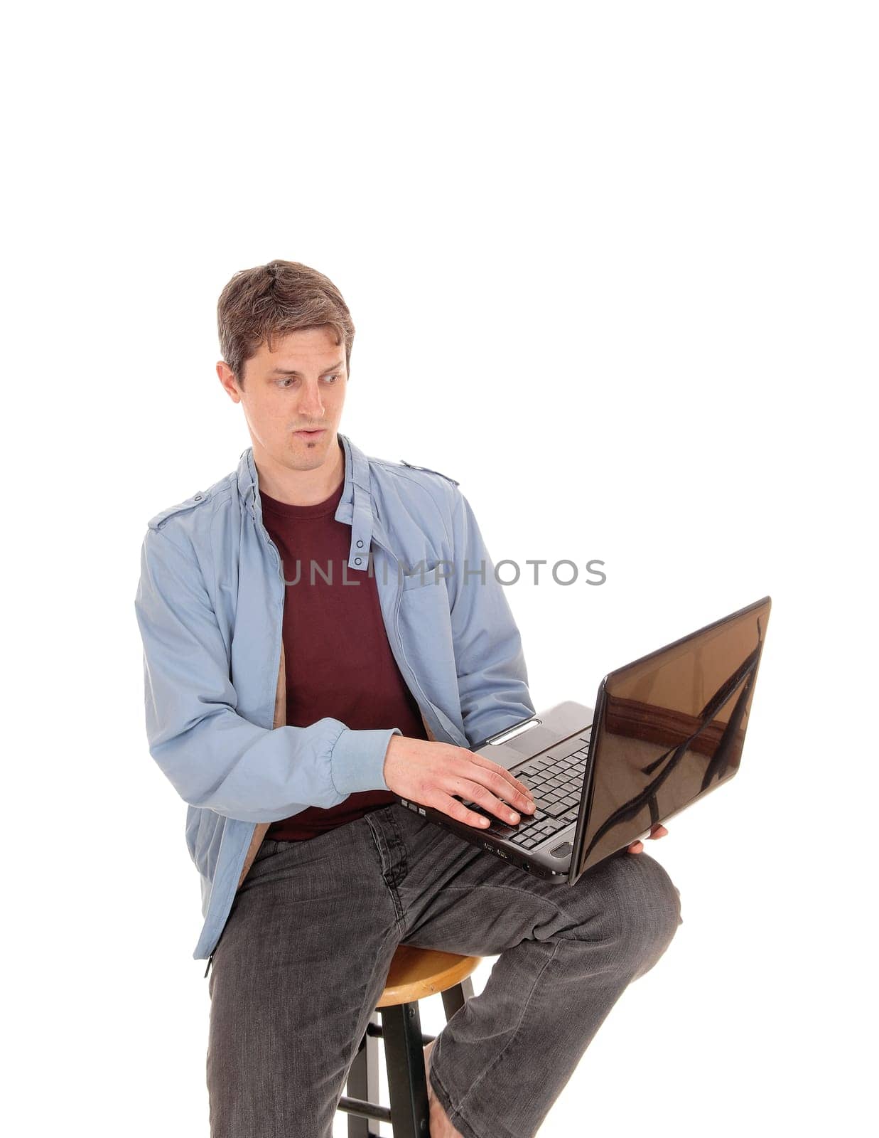 A young man sitting in a blue jacket with his laptop on his knees.looking puzzled at the screen, isolated for white background.