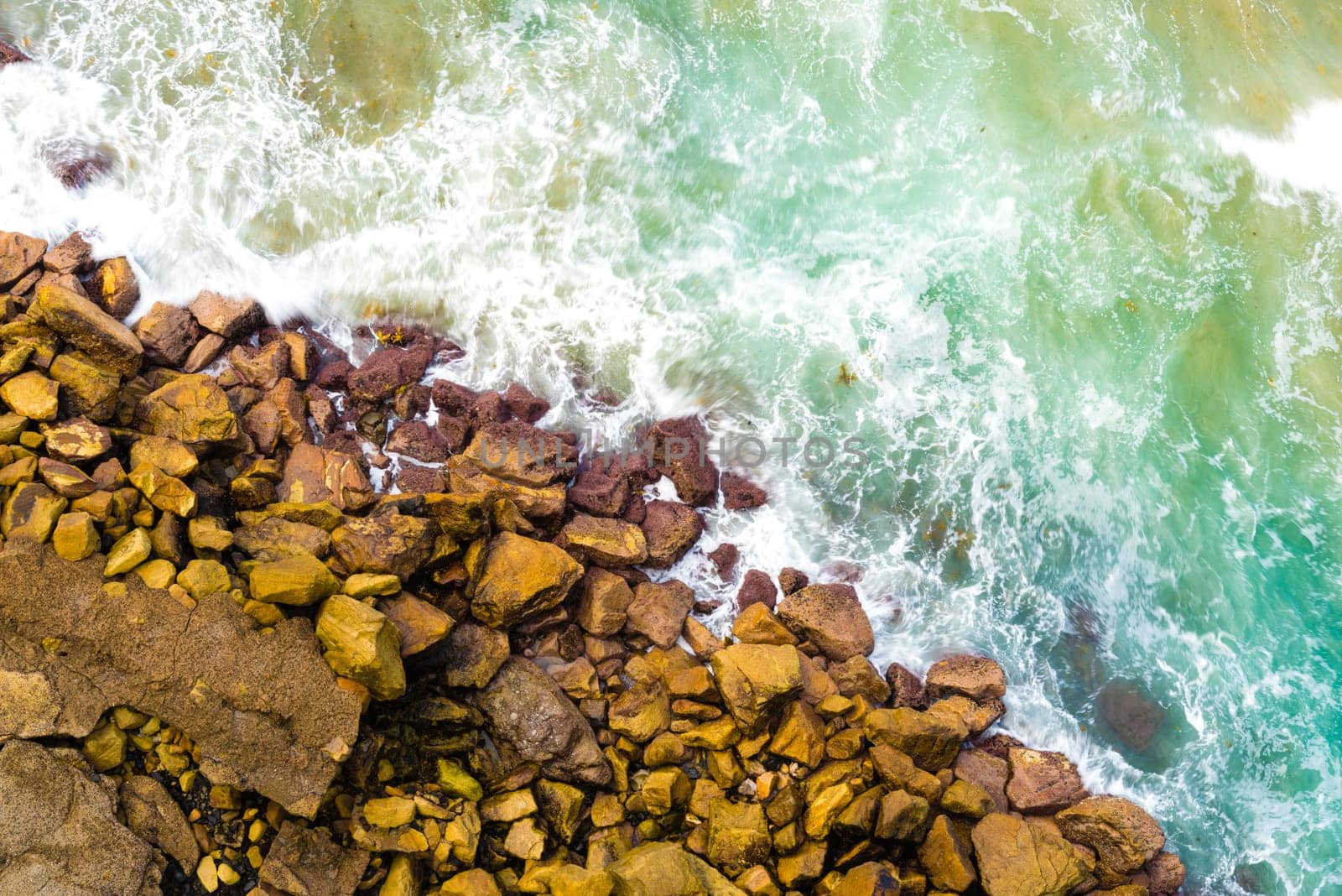 Top aerial view of blue waves crashing on rocky Australian coastline. Summer seascape with birds eye view shot over ocean waves and cliffs. Travel concept background