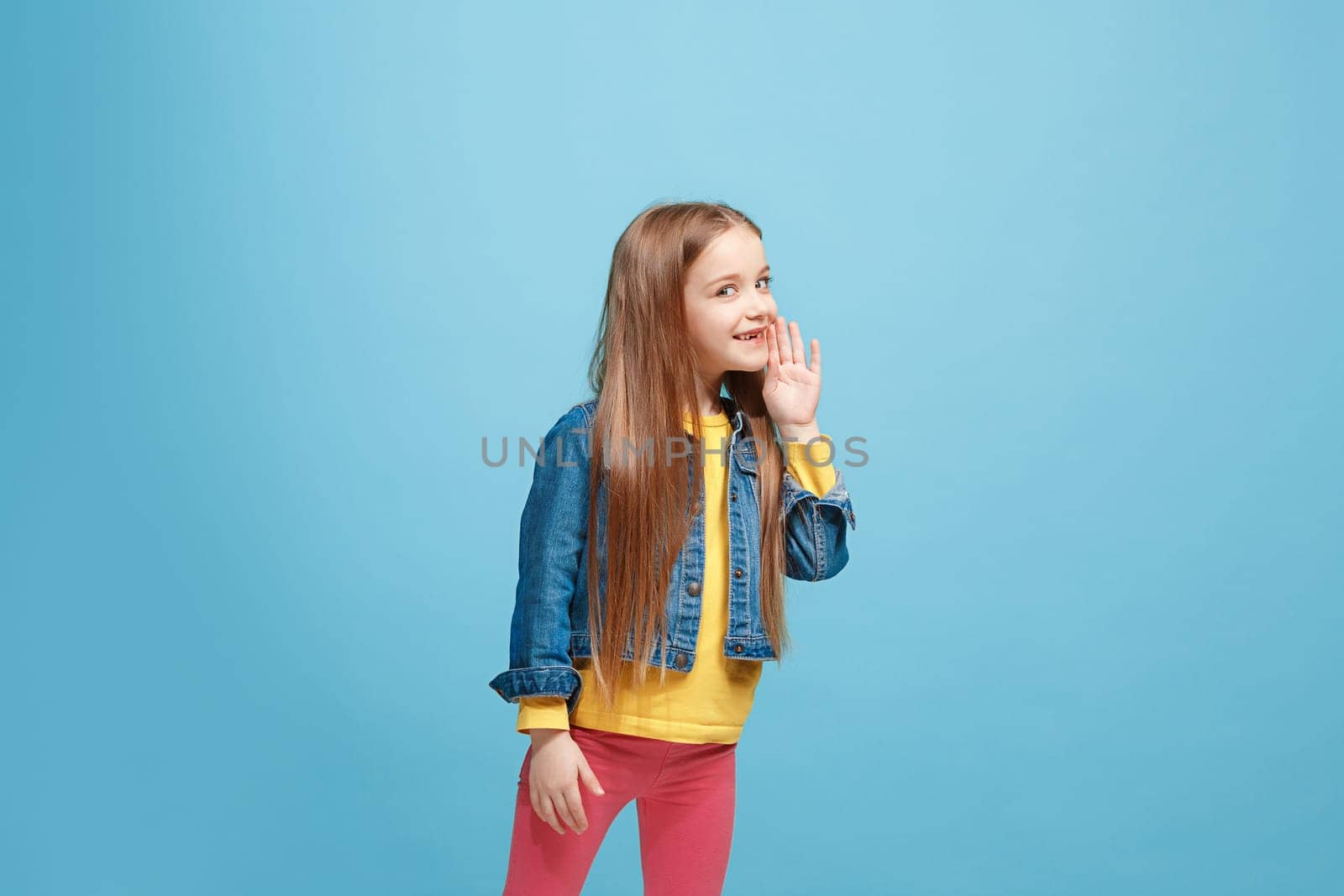 Secret, gossip concept. Young teen girl whispering a secret behind her hand isolated on trendy blue studio background. Young emotional girl. Human emotions, facial expression concept.