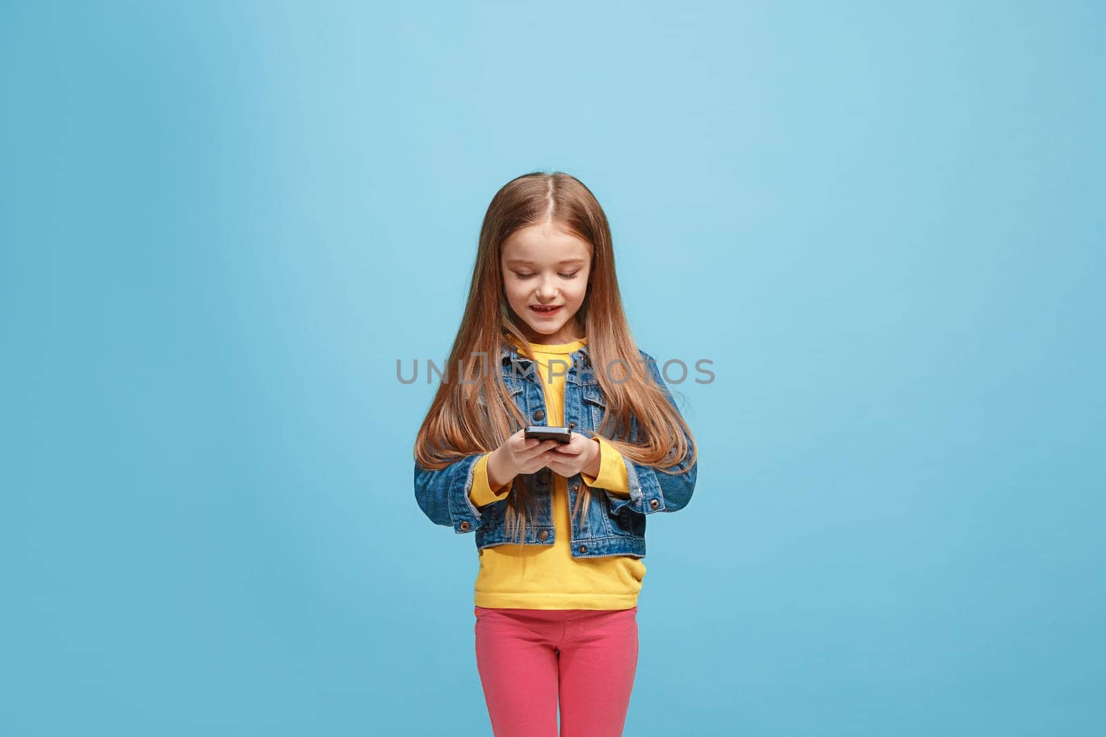 Happy teen girl standing, smiling with mobile phone over trendy blue studio background. Beautiful female half-length portrait. Human emotions, facial expression concept.