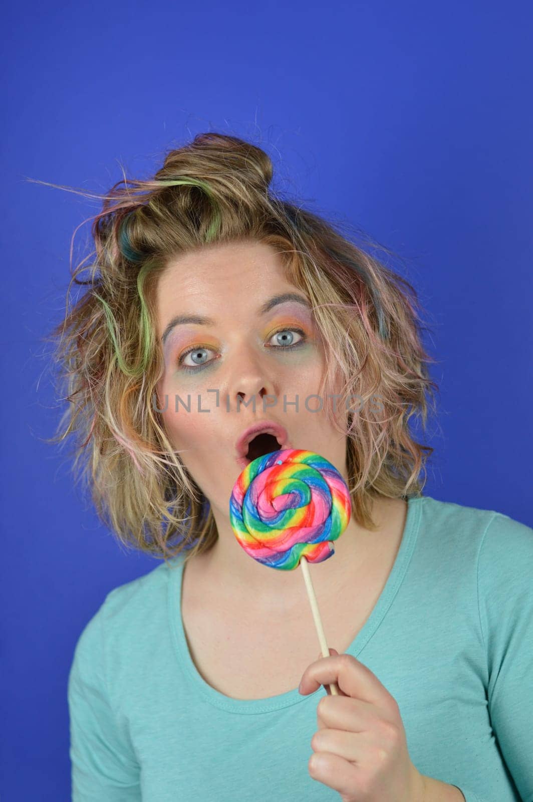 portrait of a young blond woman with colourful streaks in the hair and a big sweet multicolored lollipop in her hand looking surprised into the camera