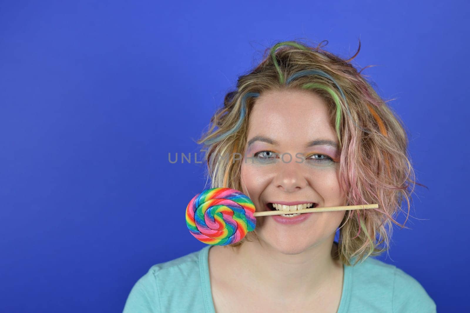 portrait of a young blond woman with colourful hair and a big lollipop between her teeth having fun and looking into the camera