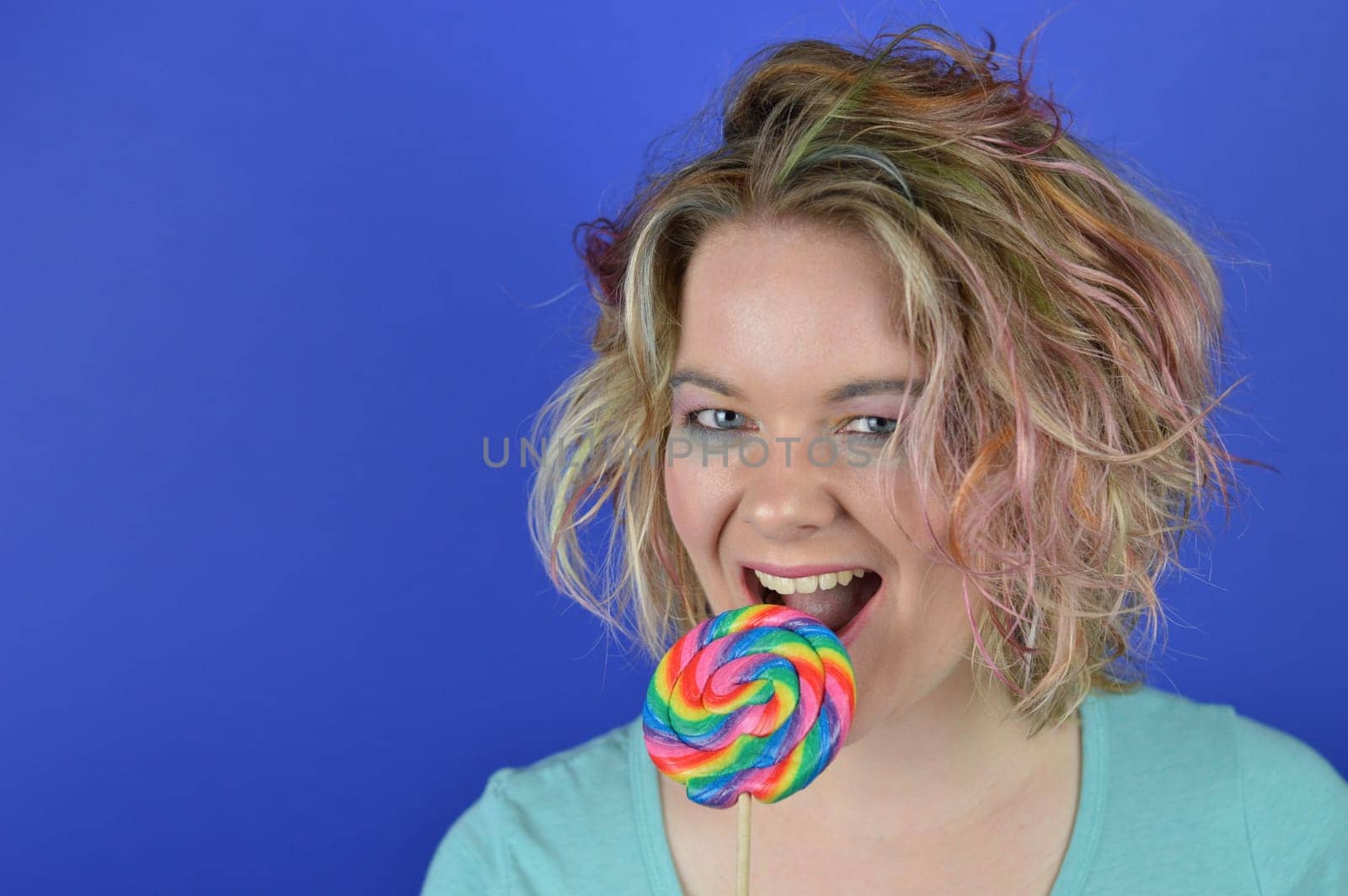 portrait of a young blond woman with colourful streaks in the hair biting into a big sweet multicolored lollipop and having fun looking into the camera