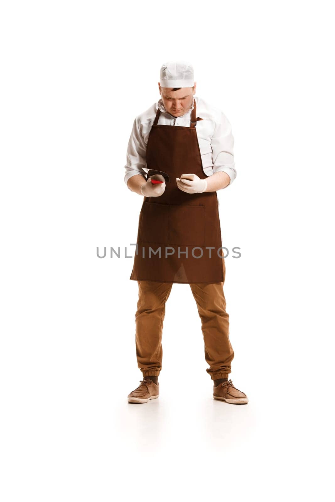 Sad butcher posing with a knife and mobile phone isolated on white studio background. The young caucasian male model in full-length. Butcher, knife, professional concept