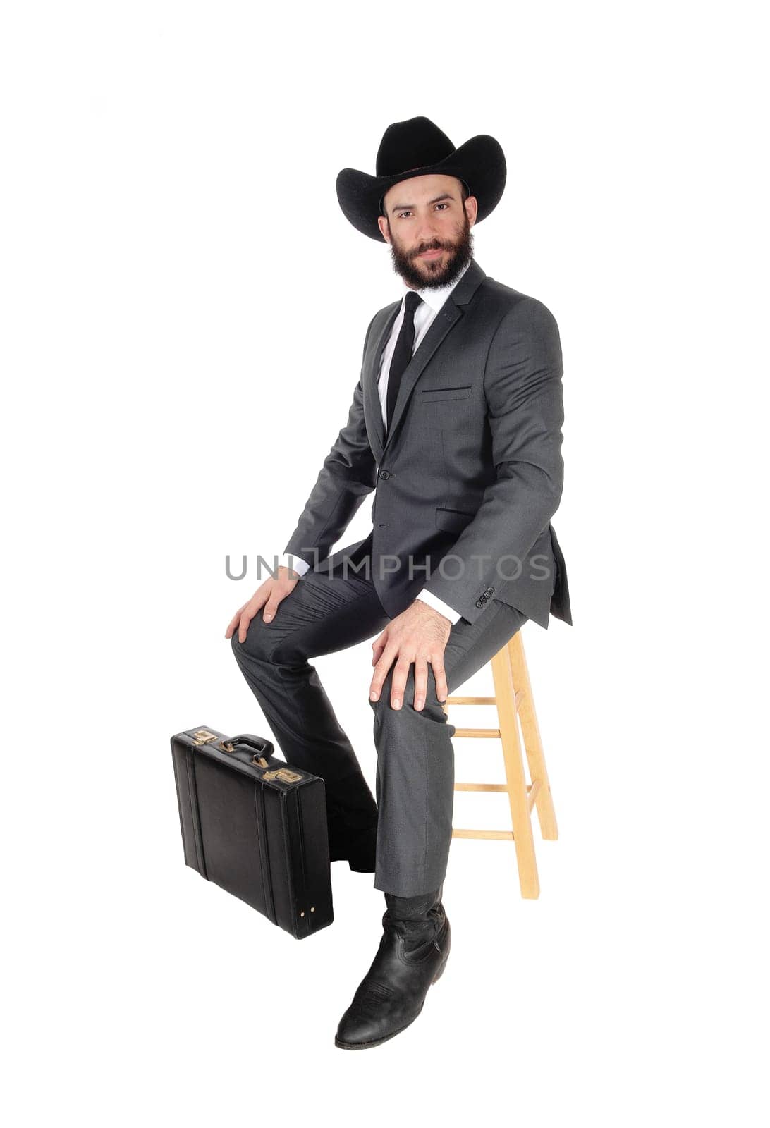 A young handsome business man wearing a suit and cowboy hat sitting.on a bar chair with his briefcase, isolated for white background.