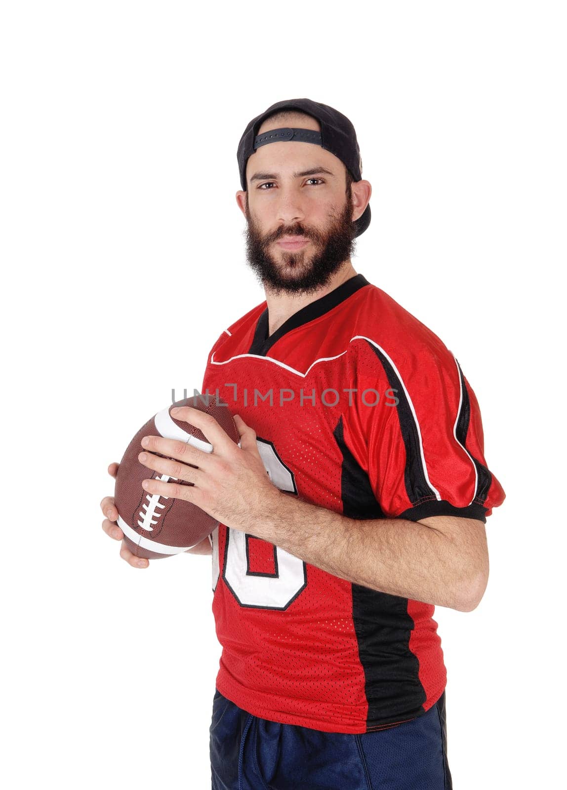 A football player in his red jersey and his brown football in his hand.standing and looking into the camera, isolated for white background.