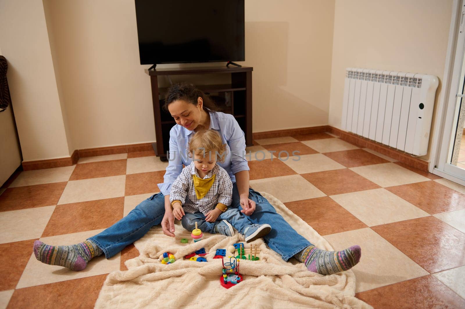 A mother and child enjoy quality time playing together on a blanket indoors. Bright toys and a cozy atmosphere portray care, happiness, and bonding.