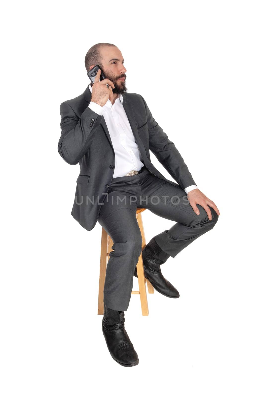 A handsome young business man sitting on a bar chair talking on his.cell phone, with a beard and suit, isolated for white background.