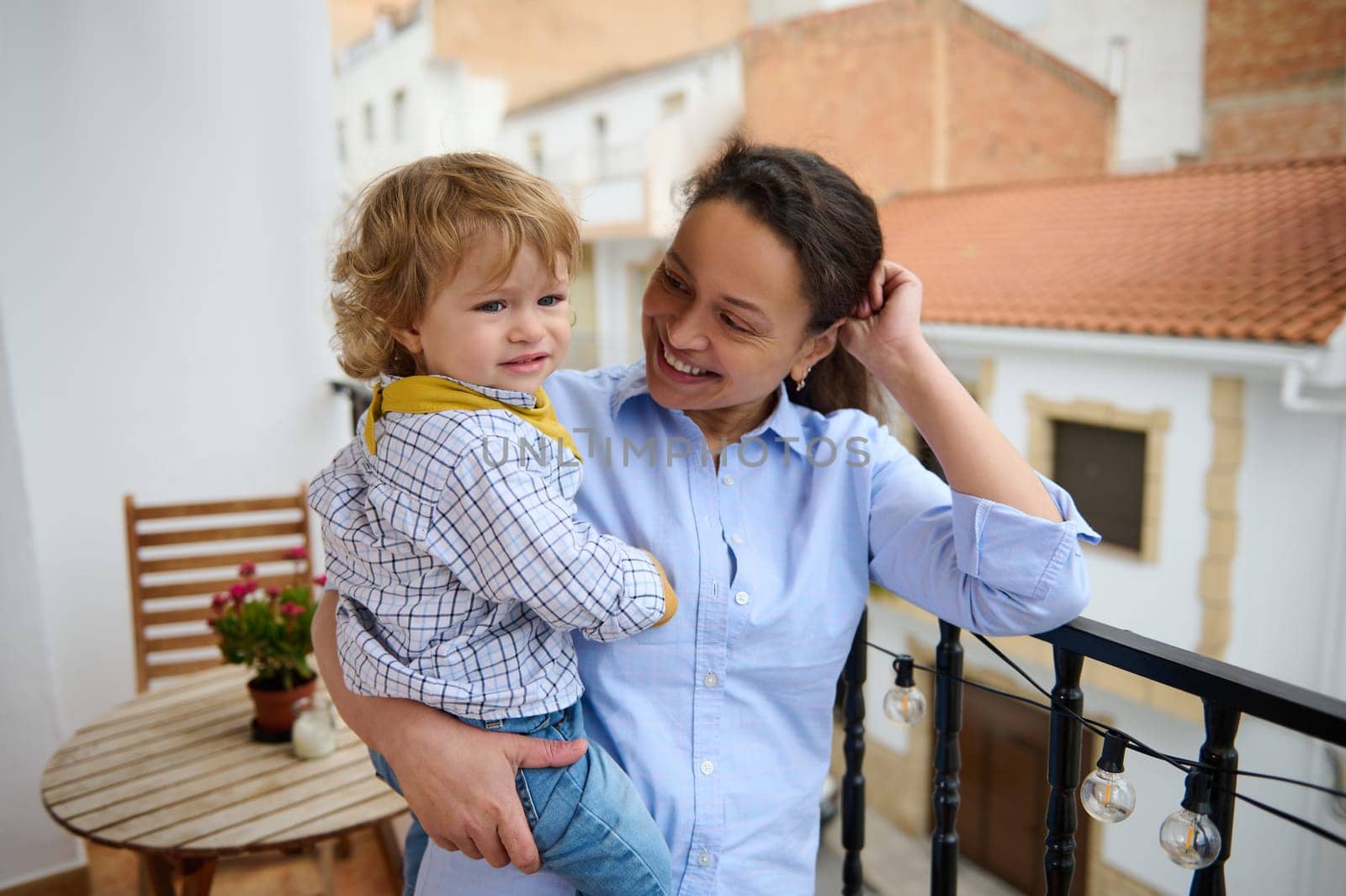 A cheerful mother enjoying maternity leave smiles as she holds her happy child on a balcony, emphasizing familial love, care, and the bond they share.