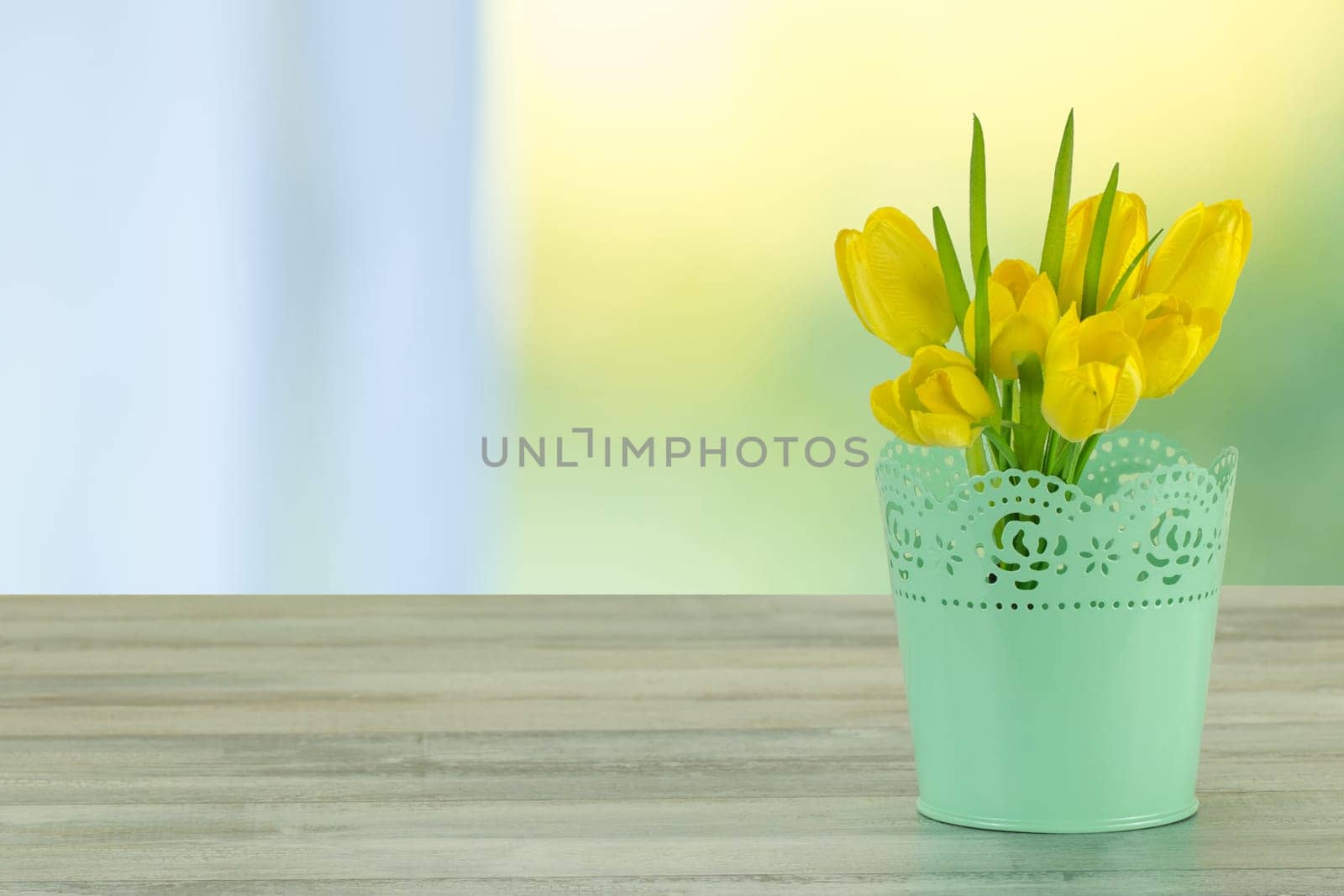 Decorative composition of yellow tulips on rustic bright wooden table. The shining bright background is symbol of spring. The bunch of yellow tulips is in decorative green vase. Empty space for your design. Card concept for mothers day, birthday, aniversary and other events.