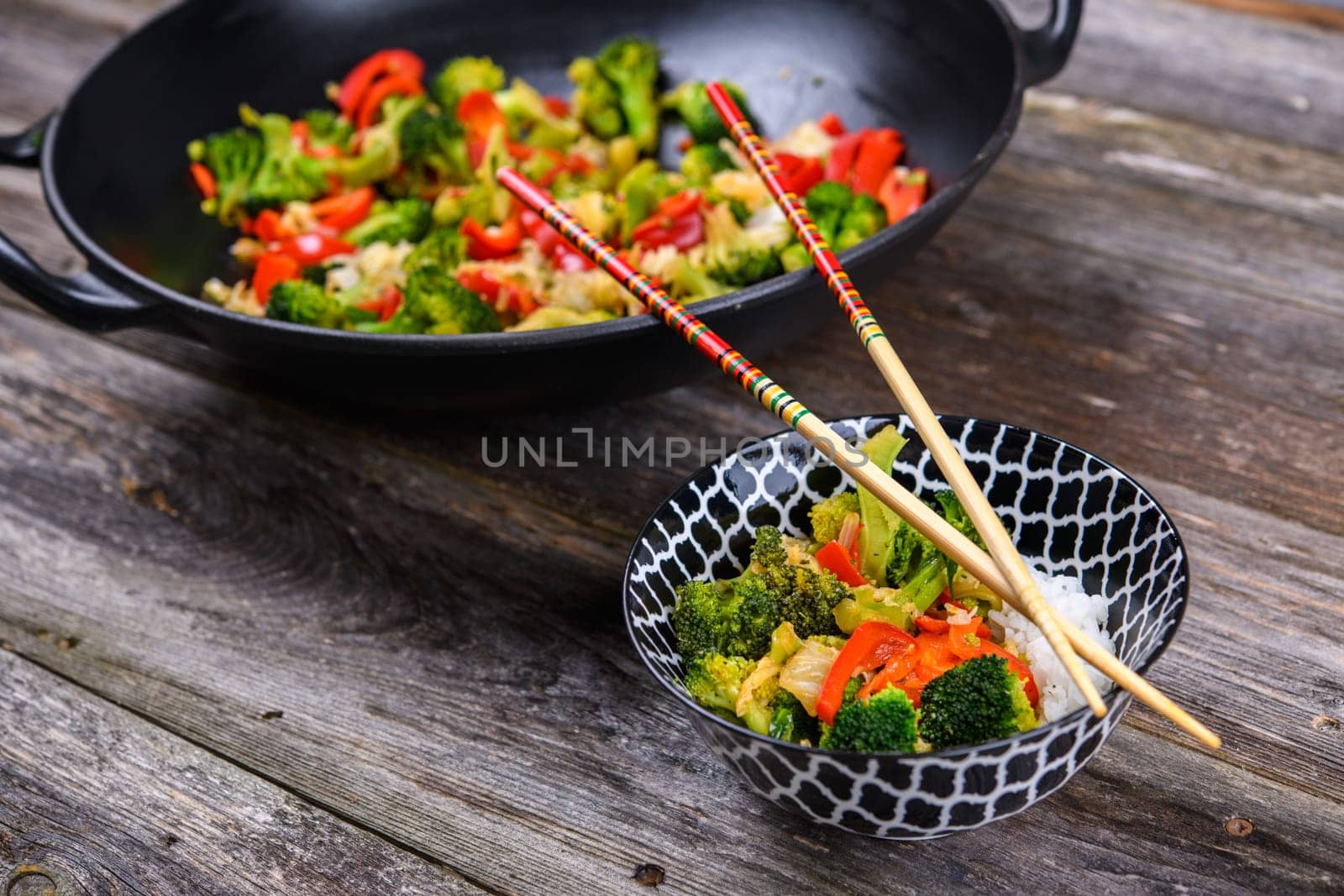 wok with colorful vegetables, bowl of rice and chopsticks on wooden ground