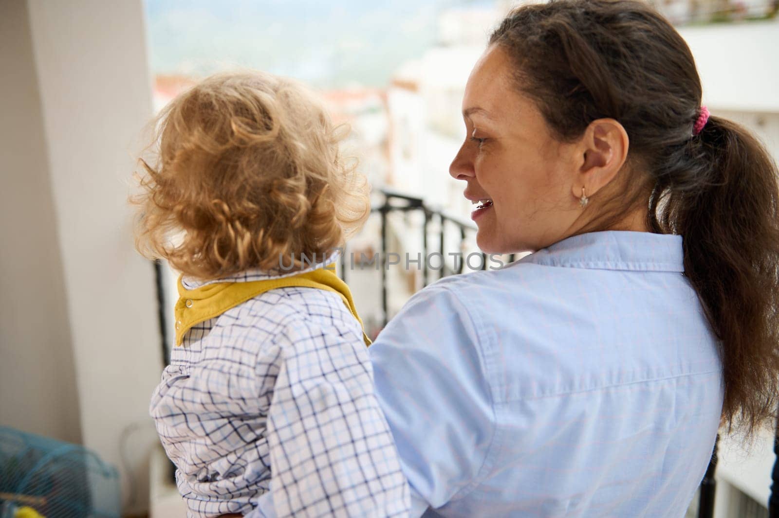 A mother holding her child stands on a balcony overlooking a bright, scenic area, creating a moment of nurturing and connection, evoking feelings of happiness and parental affection.