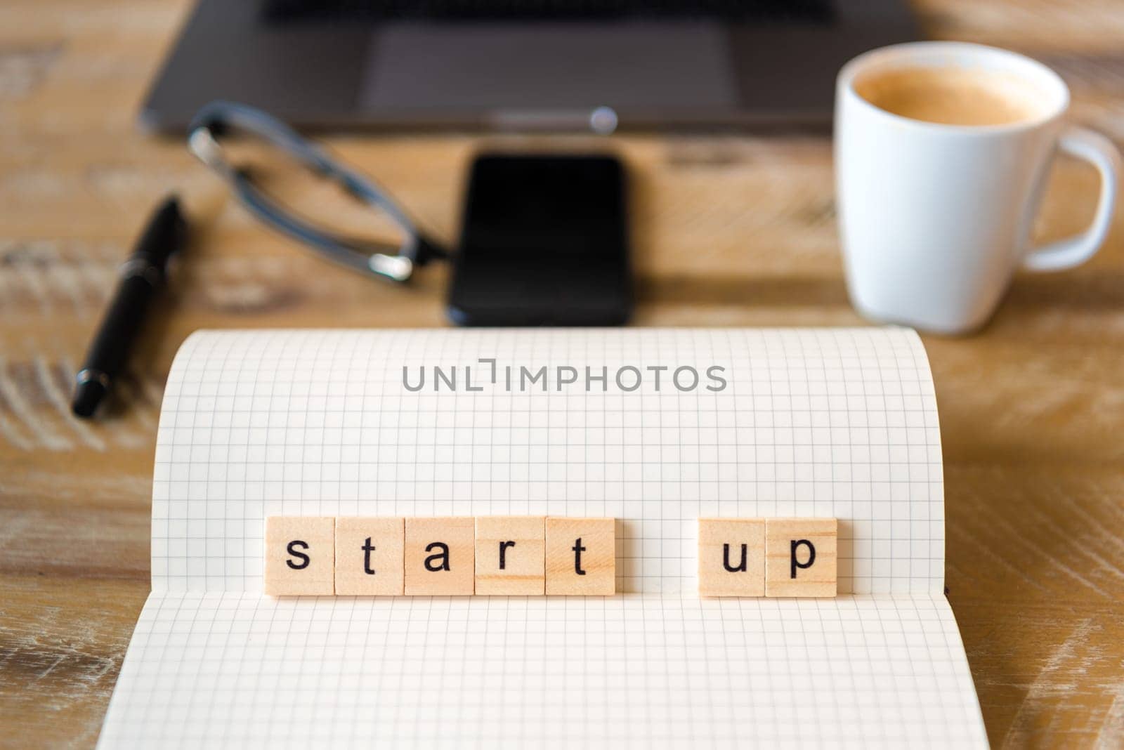 Closeup on notebook over wood table background, focus on wooden blocks with letters making Start Up word. Business concept image. Laptop, glasses, pen and mobile phone in a defocused background.