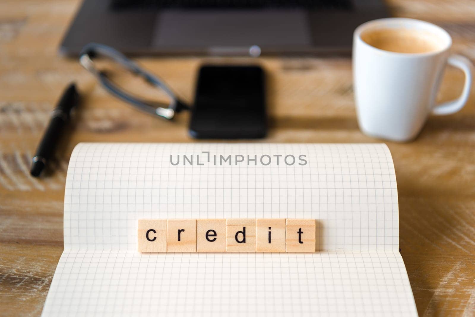 Closeup on notebook over wood table background, focus on wooden blocks with letters making Credit word. Business concept image. Laptop, glasses, pen and mobile phone in a defocused background.