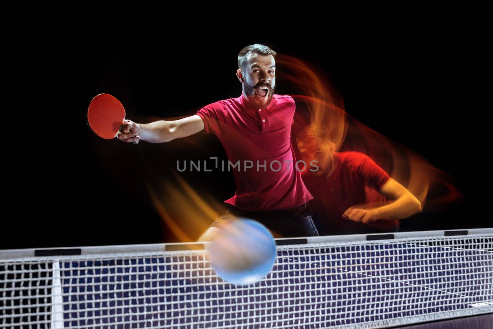 The table tennis player in motion. Fit young sports man tennis-player in play on black background with lights. Movement, sport game, stobe concepts. Professional. Human emotions, facial expression