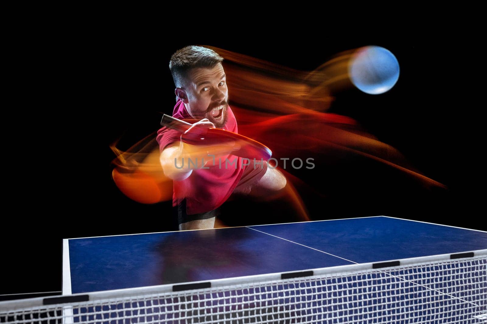The table tennis player in motion. Fit young sports man tennis-player in play on black background with lights. Movement, sport game, stobe concepts. Professional. Human emotions, facial expression