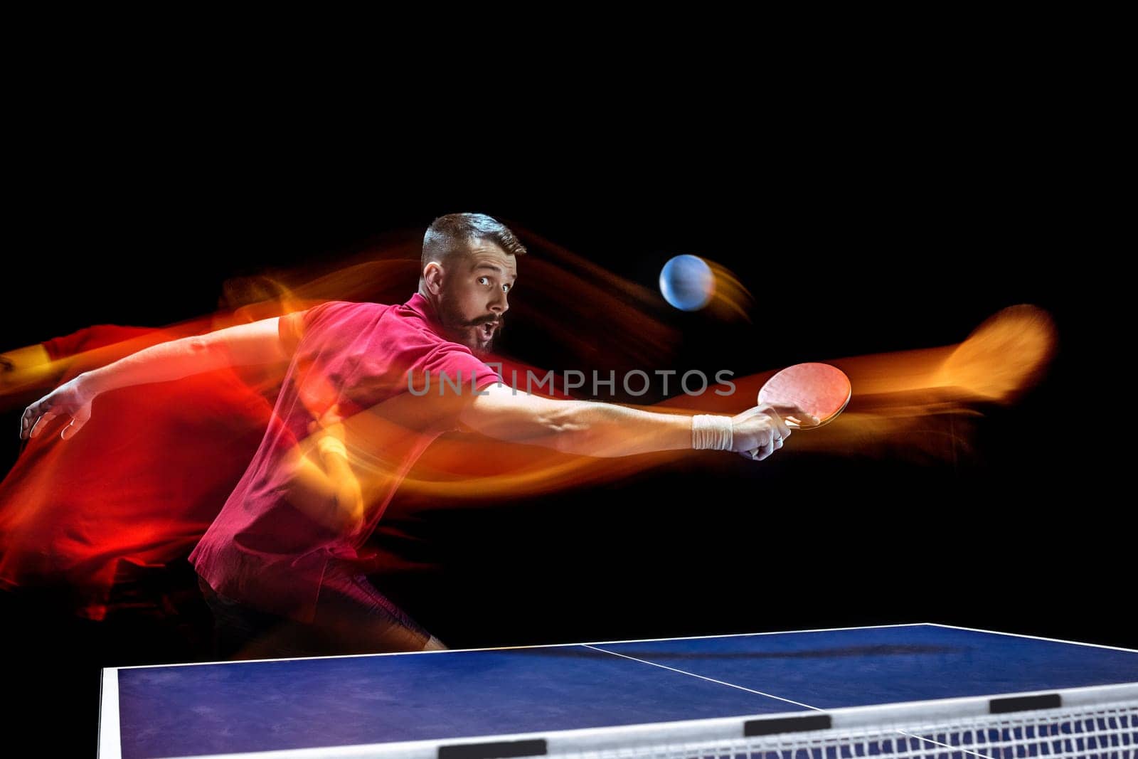 The table tennis player in motion. Fit young sports man tennis-player in play on black background with lights. Movement, sport game, stobe concepts. Professional. Human emotions, facial expression