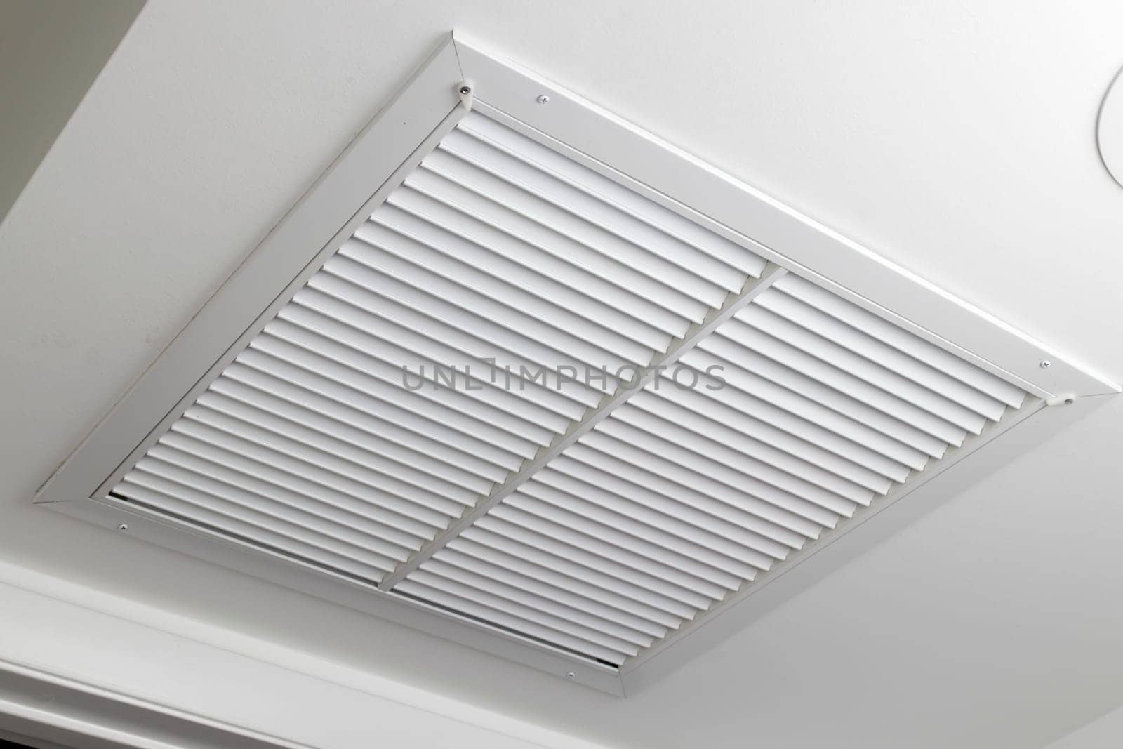 One large white painted metal furnace air vent grill with many openings on a ceiling close-up. Square outflow air filter door vent in a modern home ceiling up close.