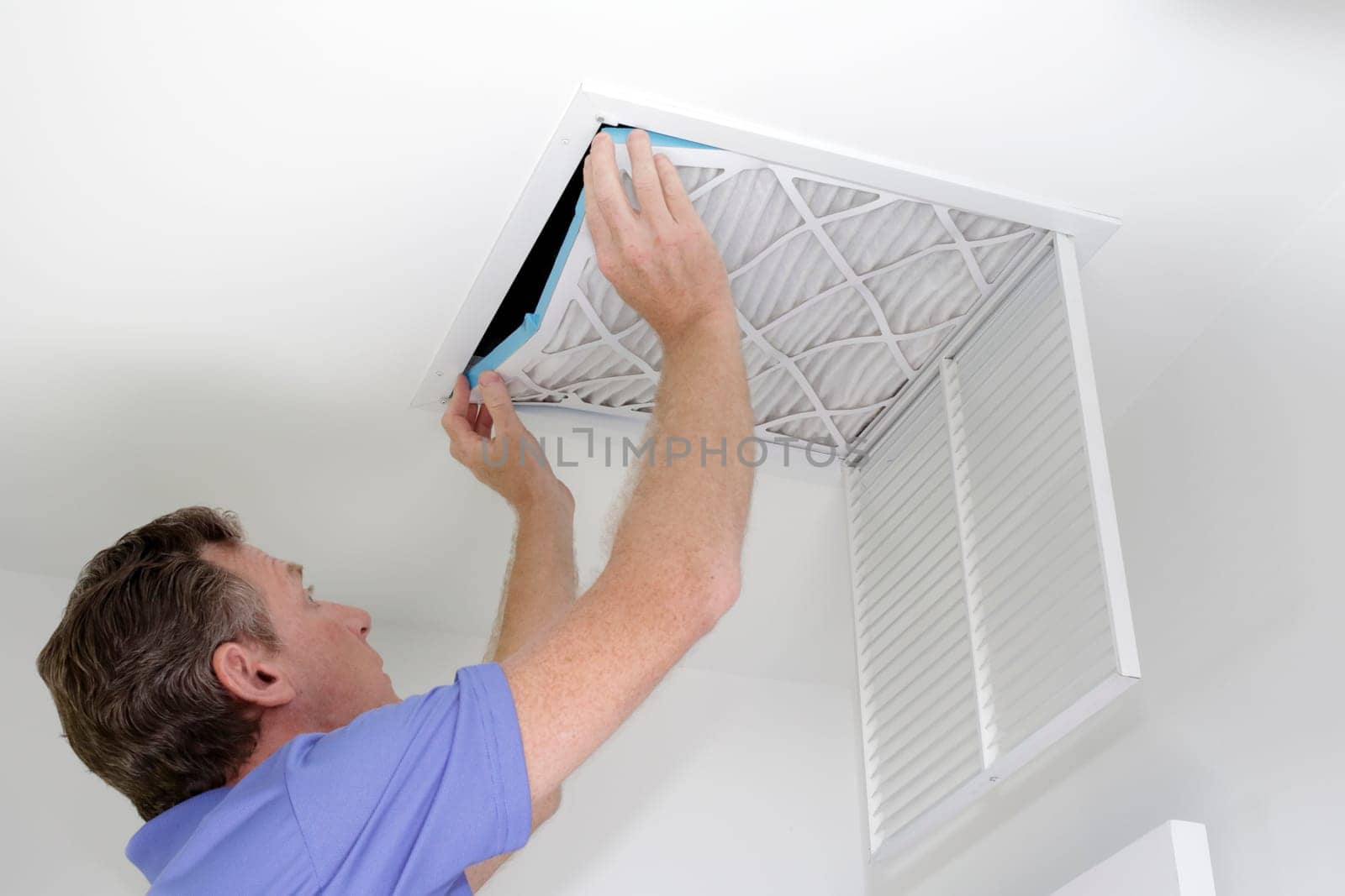 Guy placing into a square ceiling intake vent a clean white and blue air filter. As regular monthly maintenance a male puts in a new air filter in his ceiling air return.