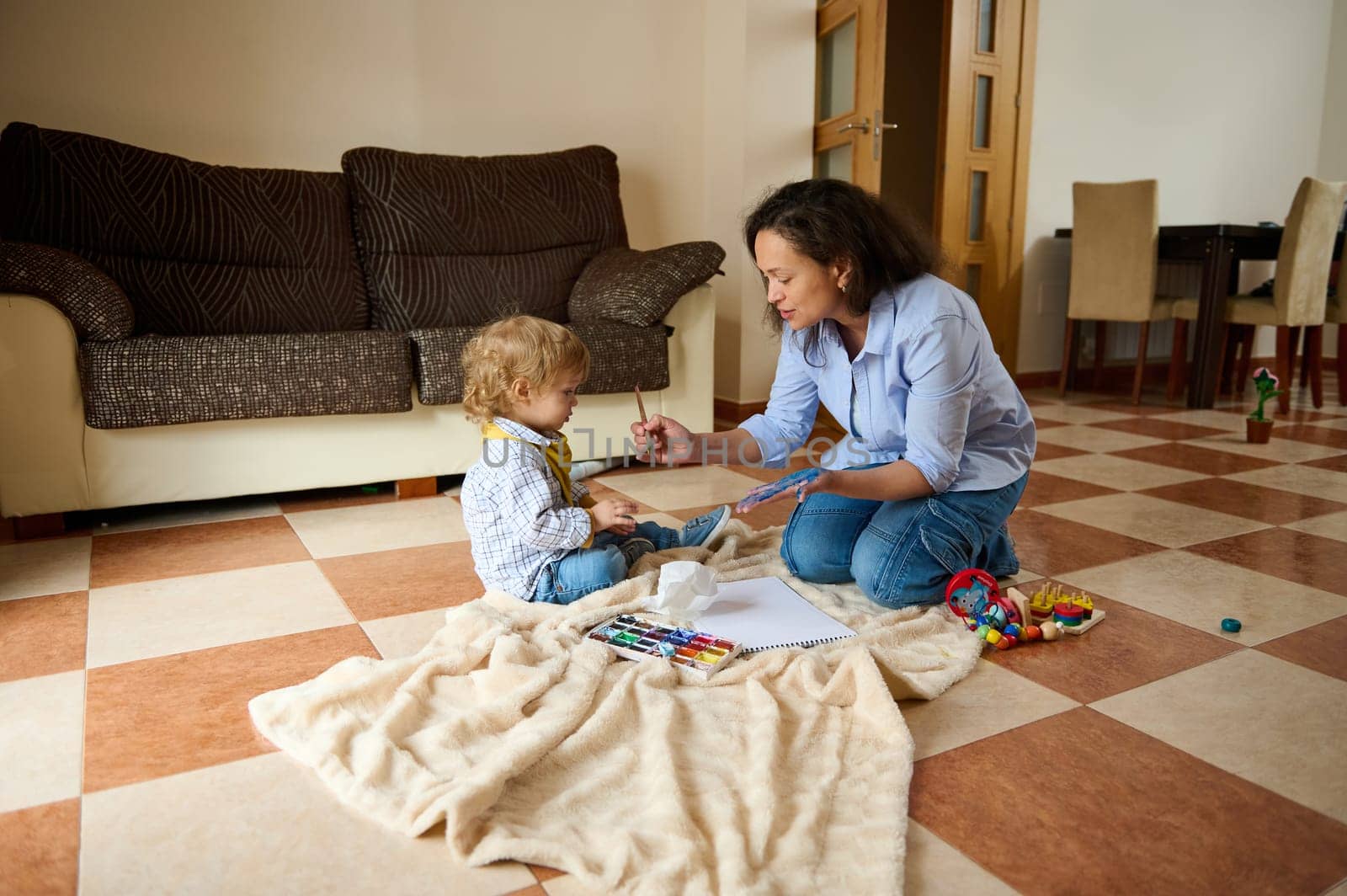 Mother joyfully engages with her toddler as they paint together in a cozy living room atmosphere, surrounded by toys, embodying the warmth of family bonding and creative interaction.
