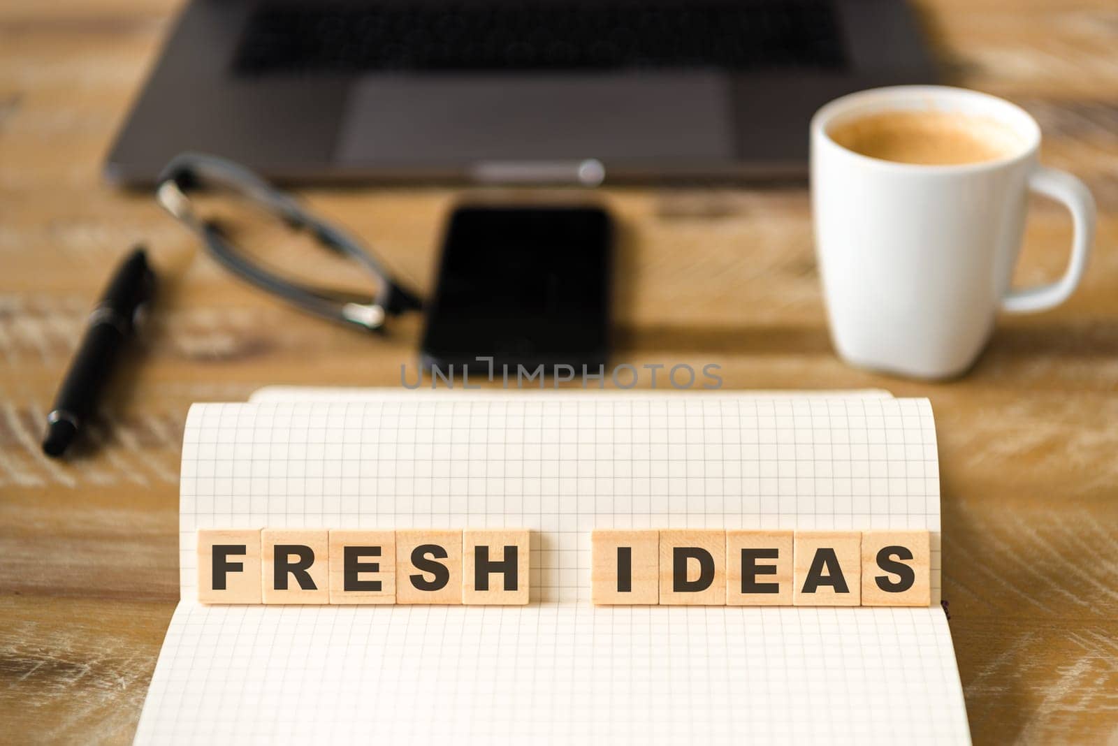 Closeup on notebook over wood table background, focus on wooden blocks with letters making Fresh Ideas text. Concept image with laptop, glasses, pen and mobile phone in defocused background