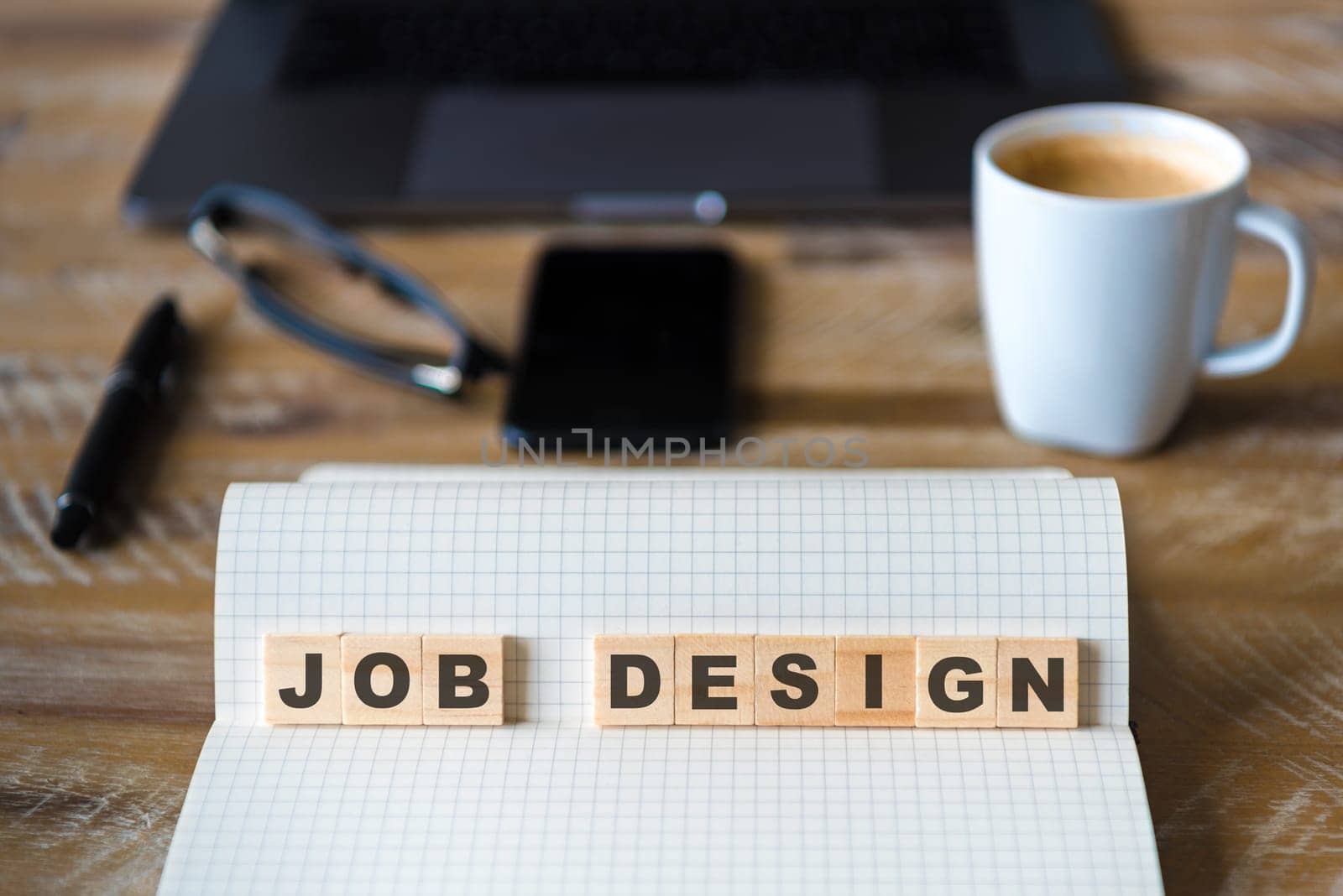 Closeup on notebook over wood table background, focus on wooden blocks with letters making JOB DESIGN words. Business concept image. Laptop, glasses, pen and mobile phone in defocused background