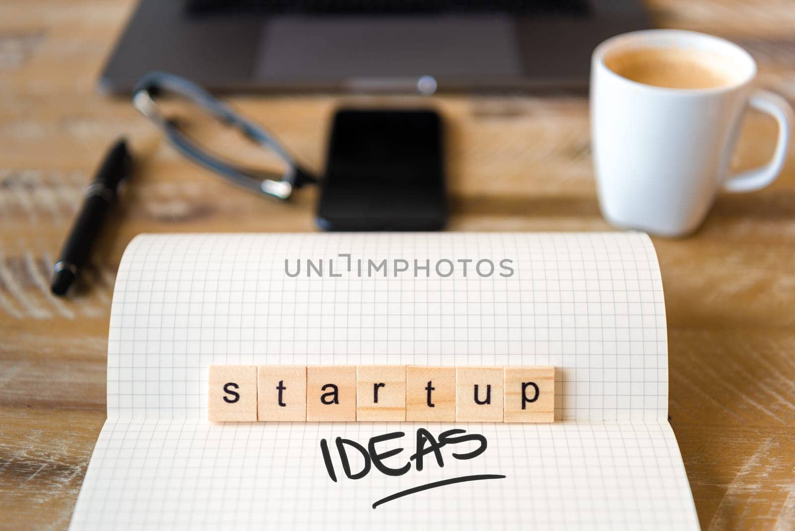 Closeup on notebook over wood table background, focus on wooden blocks with letters making Startup Ideas text. Concept image with laptop, glasses, pen and mobile phone in defocused background