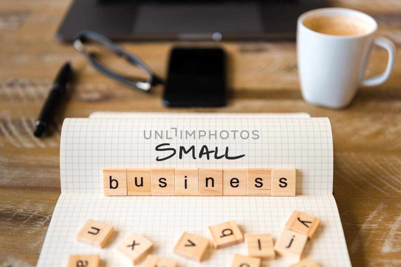 Closeup on notebook over wood table background, focus on wooden blocks with letters making Small Business text. Concept image with laptop, glasses, pen and mobile phone in defocused background
