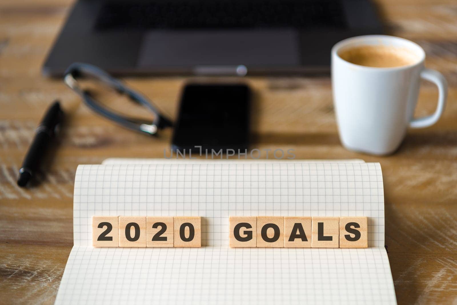 Closeup on notebook over wood table background, focus on wooden blocks with letters making 2020 Goals text. Concept image with laptop, glasses, pen and mobile phone in defocused background