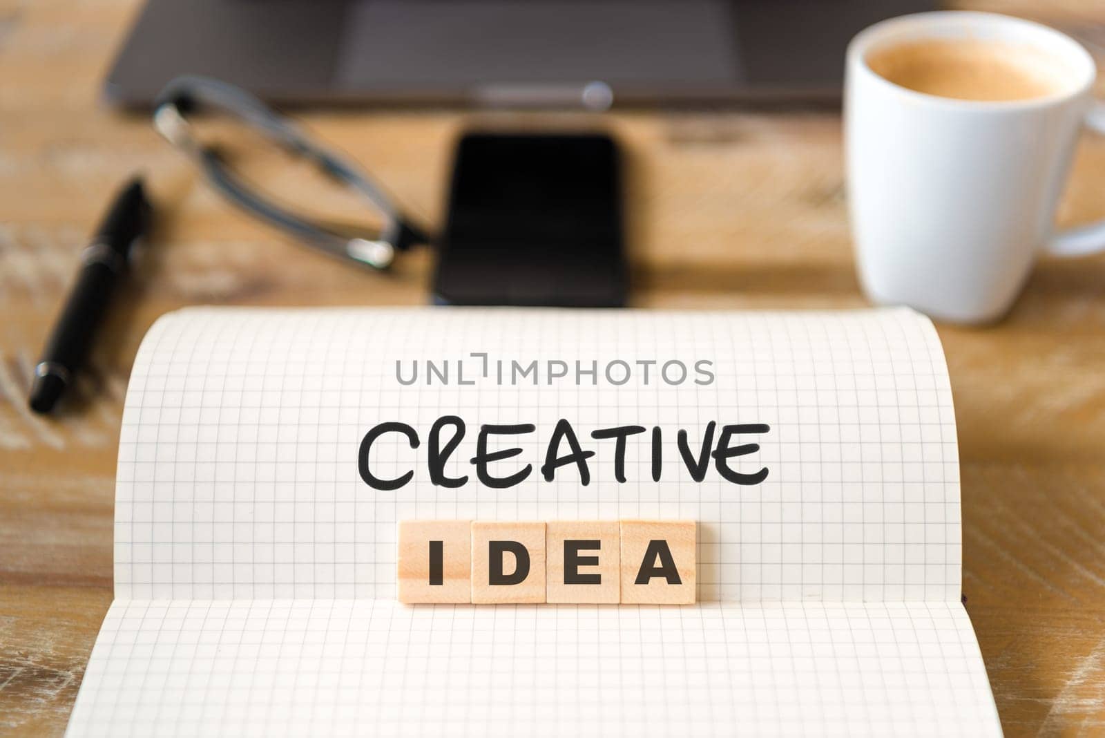 Closeup on notebook over wood table background, focus on wooden blocks with letters making Creative Idea text. Concept image with laptop, glasses, pen and mobile phone in defocused background