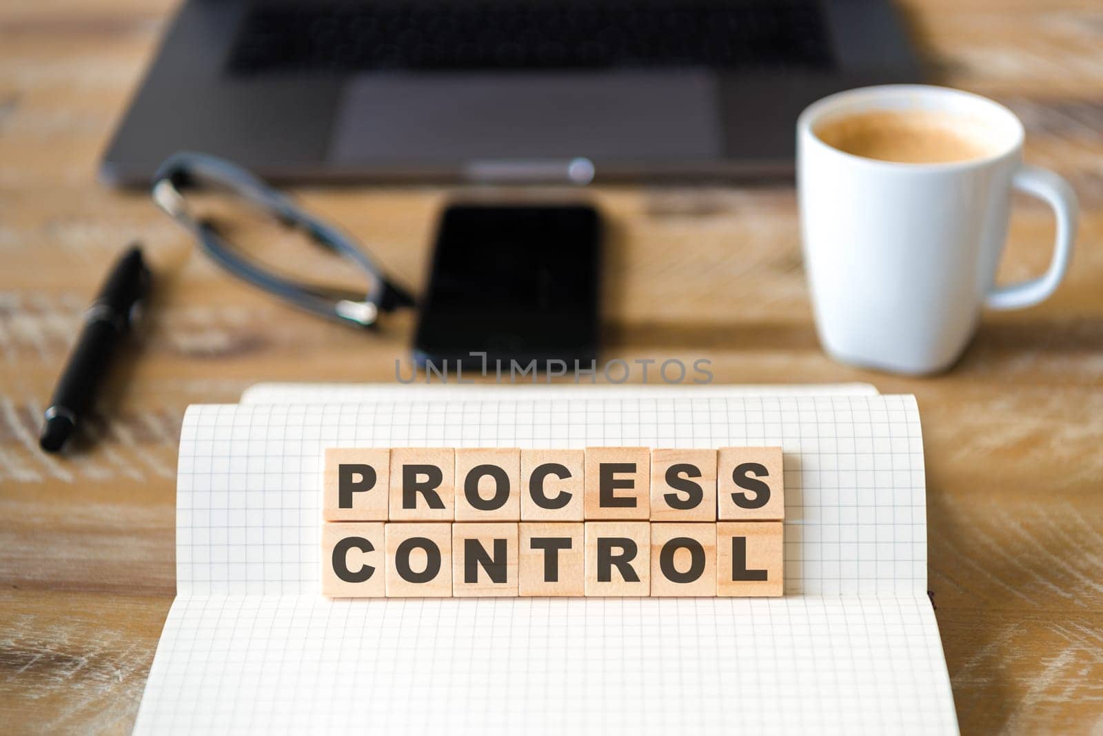 Closeup on notebook over wood table background, focus on wooden blocks with letters making PROCESS CONTROL words. Business concept image. Laptop, glasses, pen and mobile phone in defocused background