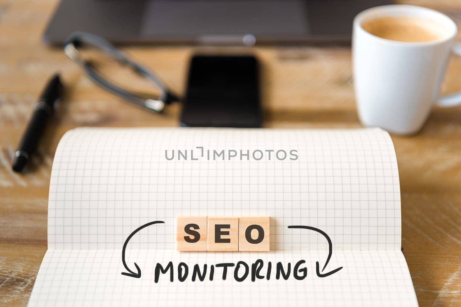 Closeup on notebook over wood table background, focus on wooden blocks with letters making SEO Monitoring text. Business concept image. Laptop, glasses, pen and mobile phone in defocused background