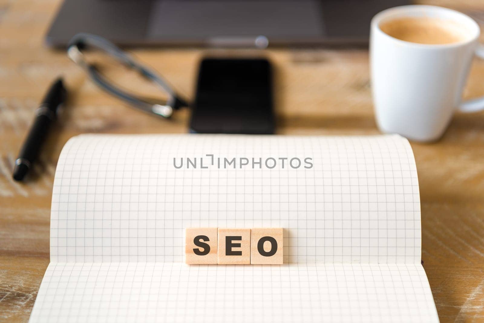 Closeup on notebook over wood table background, focus on wooden blocks with letters making SEO word. Business concept image. Laptop, glasses, pen and mobile phone in defocused background