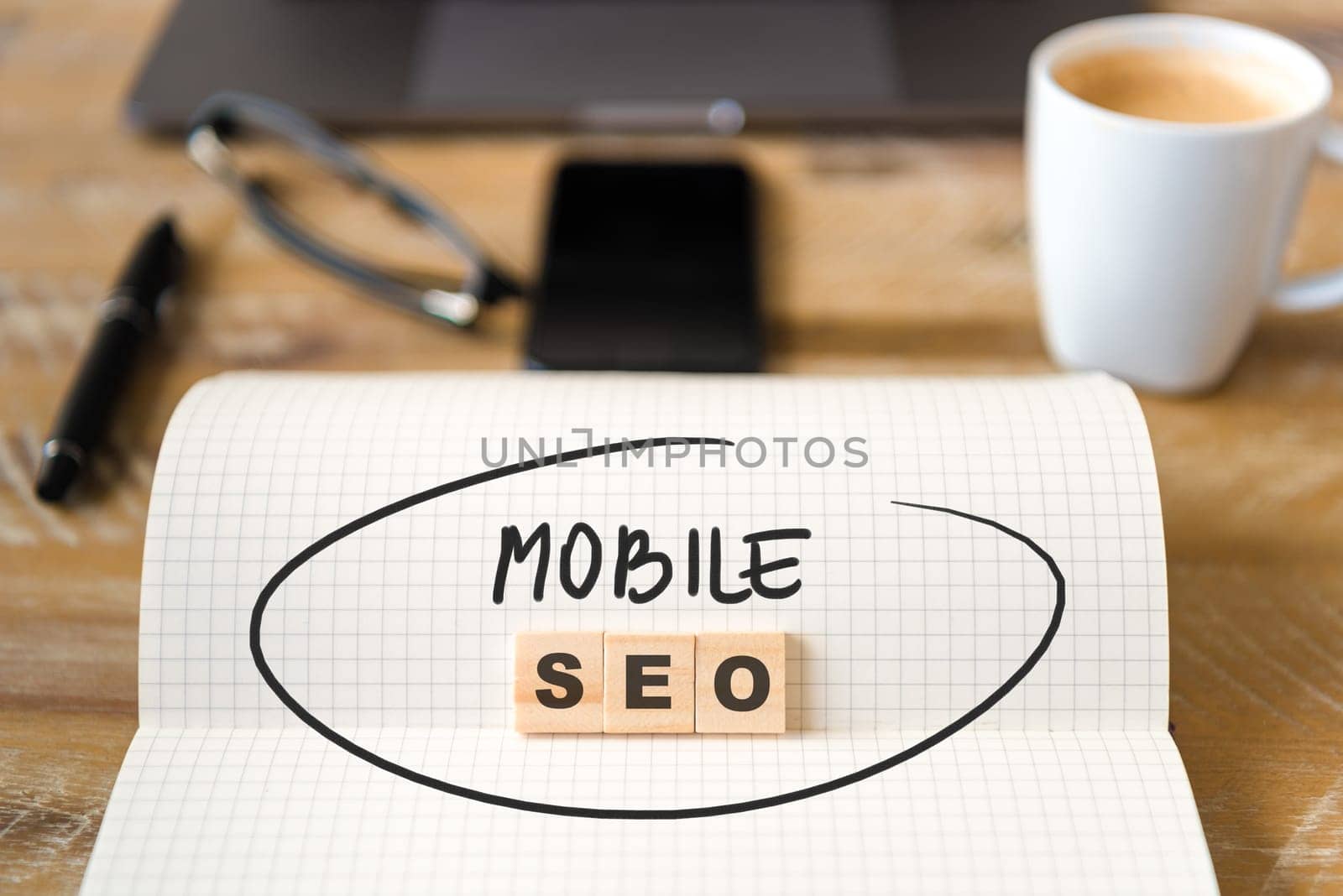 Closeup on notebook over wood table background, focus on wooden blocks with letters making Mobile SEO text. Business concept image. Laptop, glasses, pen and mobile phone in defocused background