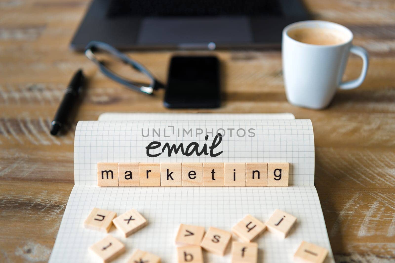 Closeup on notebook over wood table background, focus on wooden blocks with letters making Email Marketing text. Concept image with laptop, glasses, pen and mobile phone in defocused background